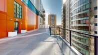 Located atop a contemporary building in the heart of Halifax, Nova Scotia, this rooftop terrace offers an impressive open-air space with sweeping views of the city skyline and Halifax Harbour. The terrace is finished with large-format grey stone pavers that create clean, defined walkways and gathering areas throughout the space. Decorative multi-coloured river rock fills the landscaped zones between pathways, adding texture and visual contrast to the overall design. Crisp white rectangular planter boxes are distributed across the terrace, providing greenery and a modern aesthetic. A bold orange and blue glass facade of the building structure anchors one side of the terrace, adding a striking architectural element and vivid colour to the space. The perimeter is lined with sleek black metal railings that ensure safety while preserving unobstructed sightlines across the surrounding neighbourhood. Views extend outward to neighbouring mid-rise and high-rise buildings, with glimpses of the Halifax waterfront visible in the distance. The terrace benefits from abundant natural light and open sky exposure throughout the day, making it a highly functional and visually dynamic outdoor space well-suited for gatherings or quiet enjoyment of the urban landscape.