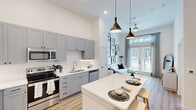 Located at 122 Sheraton Ave in Austin, TX, this thoughtfully designed one-bedroom, two-bathroom residence offers an efficient and stylish single-floor layout. The open-concept kitchen and living area serves as the heart of the home, showcasing soaring ceilings that amplify the sense of space. The kitchen is outfitted with gray shaker-style cabinetry extending to ceiling height, a white geometric tile backsplash, white quartz countertops, and a full suite of stainless steel appliances including a refrigerator, electric range, built-in microwave, and dishwasher. A central kitchen island with pendant lighting above provides additional prep and dining space. Light wood-tone flooring runs throughout, complementing the bright, white walls. The living area benefits from large French doors that invite abundant natural light and outdoor views. The bedroom is a serene retreat, featuring a large window with sheer curtains, views of the surrounding greenery, and direct access to a well-appointed bathroom with a walk-in closet. The closet features built-in shelving and dual hanging rods for ample storage. Both bathrooms are finished with clean, modern fixtures, a frameless mirror, and white vanities. The overall ambiance is clean, contemporary, and efficiently planned.