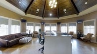This distinctive single-floor space immediately captures attention with its octagonal architectural design and dramatically vaulted ceiling. The ceiling is a standout feature, clad in rich, dark wood planking and supported by exposed wooden beams that converge toward the center, where an elegant chandelier serves as the focal point. The elevated ceiling height lends an open, grand atmosphere to the interior.

Abundant natural light floods the space through a series of large windows that line the perimeter walls, each fitted with tailored Roman shades that offer a clean, polished look. Multiple flat-screen monitors are wall-mounted at intervals around the room, suggesting a functional, client-facing purpose.

At the heart of the space sits a prominent white reception or service desk, designed to accommodate staff and facilitate visitor interactions. The polished concrete flooring adds a modern, low-maintenance element that complements the warm wood tones overhead.

A comfortable seating area is arranged near one side of the room, featuring upholstered armchairs, a tufted ottoman, and a decorative side table with a lamp and floral arrangement, creating a welcoming lounge atmosphere. The overall ambiance balances professional functionality with warm, inviting design elements, making it well-suited as a leasing office or community welcome center.