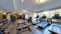 This fitness center offers a spacious, single-floor layout thoughtfully designed to accommodate a comprehensive range of workout options. The space is divided into functional zones, each dedicated to a specific type of exercise, creating an organized and efficient environment for users of all fitness levels.

The cardio zone is prominently positioned along a wall of large, wood-framed windows that flood the area with natural light and provide pleasant views of surrounding greenery. Multiple treadmills, elliptical machines, stair climbers, and stationary and spin bikes are arranged to maximize both space and sightlines.

The strength training area features a multi-station cable machine and a dedicated free weights section, complete with a well-stocked rack of dumbbells and resistance tools including TRX suspension trainers and medicine balls. Adjustable weight benches and a rowing machine round out the equipment offerings, ensuring a full-body workout capability.

Underfoot, durable rubber flooring provides cushioning and traction throughout the space. Overhead, a combination of recessed lighting, flush-mount ceiling fixtures, and track lighting ensures the entire facility is well-lit at all times. The neutral-toned walls and clean architectural lines contribute to a modern, focused atmosphere. A dedicated entry door provides convenient access, making this fitness center a highly functional and well-appointed amenity.