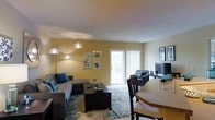This single-floor space features an open-concept layout that seamlessly connects the living and dining areas, creating a cohesive and functional environment. The living area is anchored by a large gray sectional sofa accented with teal throw pillows, complemented by a dark wood coffee table resting on a patterned area rug. A multi-arm arc floor lamp provides ambient lighting, enhancing the warm yellow tones of the walls throughout the space.

A sliding glass door with vertical blinds fills the living area with natural light and provides direct access to an outdoor balcony, offering views of the surrounding exterior. Adjacent to the living space, a media setup includes a flat-screen television and coordinating accent chairs in a teal and gray palette, maintaining a consistent design aesthetic.

The dining area transitions naturally from the living space and features a wood-toned table paired with dark wood chairs, suitable for everyday meals or entertaining. The neutral carpeted flooring ties both zones together, while the overall layout maximizes the sense of openness and flow throughout the single-level floor plan.