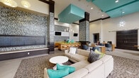 Located at 7951 NE Bayshore Ct in Miami, FL, this single-story clubhouse presents a sleek, modern design characterized by soaring double-height ceilings and floor-to-ceiling windows that flood the interior with natural light while offering views of the surrounding tropical landscape and pool area. The open-plan layout is anchored by a striking teal ceiling feature element suspended above the central lounge, where generously sized neutral sofas are accented with vibrant teal and lime green throw pillows, creating an inviting social hub. Tall mosaic-tiled accent columns with a linear, textured pattern add architectural drama throughout the space, complementing the patterned carpet underfoot. A lime green countertop coffee and refreshment bar is equipped with a Lavazza coffee machine, additional brewing stations, and under-counter appliances, backed by the same signature mosaic tile wall. Dedicated workstation areas with modern task chairs and wood-toned desks provide functional workspace, while a billiards table offers recreational options. Built-in bench seating with upholstered panels lines a tiled alcove near the entrance corridor. Large-format tile flooring extends through transitional corridors, and multiple wall-mounted flat-screen televisions are positioned throughout. The overall ambiance is polished, energetic, and distinctly Miami-modern.