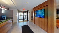 Located at 5 Dennis Street in New Brunswick, NJ, this single-floor lounge offers a thoughtfully designed communal space that combines functional and aesthetic elements. The entrance features glass doors that allow natural light to flood the foyer, which is finished with large-format tile flooring and a wall-mounted digital display screen set against warm wood paneling—ideal for broadcasting information and announcements.

Moving inward, a streamlined reception desk crafted from warm wood tones serves as a central focal point, equipped with multiple computer workstations and accented by a suspended linear light fixture above. A textured accent wall in muted green adds visual depth to the reception area.

The lounge area extends further into the space, showcasing a vibrant orange sectional sofa complemented by bold abstract artwork and a tall floor lamp. A secondary seating alcove features rounded, pod-style chairs in deep plum and charcoal tones arranged around a low glass coffee table, with a window offering views to the exterior streetscape. Throughout, light-toned tile flooring and recessed lighting create a clean, cohesive atmosphere that balances modern design with comfort.