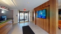 Located at 5 Dennis Street in New Brunswick, NJ, this single-floor lounge offers a thoughtfully designed communal space that combines functional and aesthetic elements. The entrance features glass doors that allow natural light to flood the foyer, which is finished with large-format tile flooring and a wall-mounted digital display screen set against warm wood paneling—ideal for broadcasting information and announcements.

Moving inward, a streamlined reception desk crafted from warm wood tones serves as a central focal point, equipped with multiple computer workstations and accented by a suspended linear light fixture above. A textured accent wall in muted green adds visual depth to the reception area.

The lounge area extends further into the space, showcasing a vibrant orange sectional sofa complemented by bold abstract artwork and a tall floor lamp. A secondary seating alcove features rounded, pod-style chairs in deep plum and charcoal tones arranged around a low glass coffee table, with a window offering views to the exterior streetscape. Throughout, light-toned tile flooring and recessed lighting create a clean, cohesive atmosphere that balances modern design with comfort.