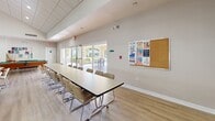 Located at 20891 NW Ninth Ct in Miami Gardens, FL, this single-floor community facility offers a generous and versatile layout designed to support a variety of group activities and social gatherings. The main multipurpose room is the heart of the space, featuring soaring vaulted ceilings, warm luxury vinyl plank flooring, and an abundance of natural light streaming in through large sliding glass doors that open to an outdoor patio area. Long conference-style tables with cushioned chairs provide ample seating for meetings and events, while a bulletin board keeps residents informed of community activities. A full-size billiards table anchors one end of the room, adding a recreational focal point. A cozy lounge area with a television, bookshelves, and comfortable seating offers a relaxed retreat, alongside a dedicated computer workstation area. The well-appointed kitchen features rich dark wood cabinetry, granite countertops, a large central island with pendant lighting, stainless steel appliances, and light wood-tone flooring. The exercise room is outfitted with a stationary bike, a full dumbbell rack, balance equipment, and rubberized flooring, providing a practical space for fitness. Recessed and drop-panel lighting throughout ensures a bright, welcoming ambiance across the entire facility.