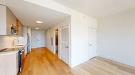 Located at 166 Van Reipen Avenue in Jersey City, NJ, this contemporary single-floor residence offers a well-designed layout comprising a kitchen, living room, and bathroom. Light oak hardwood flooring flows throughout the unit, complementing the clean, modern aesthetic and bright white walls.

The galley-style kitchen is outfitted with stainless steel appliances, including a full-size refrigerator, electric range, built-in microwave, and dishwasher. Cabinetry combines white upper cabinets with warm wood-toned lower units and open shelving, accented by white tile backsplash and light quartz countertops. The kitchen opens directly into the living area, creating an efficient, connected layout.

The living room benefits from a large floor-to-ceiling window with black framing, flooding the space with natural light and offering elevated panoramic views of the Jersey City skyline and surrounding urban landscape. A wall-mounted HVAC unit provides climate control.

The bathroom features a full soaking tub with shower, finished with large-format neutral beige wall tiles. A floating navy blue vanity with a white countertop, undermount sink, and brushed nickel fixtures adds a modern design accent, while an illuminated mirror enhances the clean, bright atmosphere. The space is completed with light tile flooring throughout the bathroom.