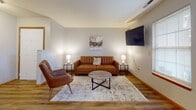 Located at 304 Turnberry Dr in Bloomington, IL, this single-floor residence offers a well-organized and functional layout designed for comfortable everyday living. The living room is a welcoming space finished with warm hardwood-style flooring, neutral gray walls, and a wall-mounted flat-screen TV. A tufted leather sofa, accent chair, and a round coffee table atop a patterned area rug create an inviting seating arrangement, complemented by natural light streaming through a window with blinds.

The kitchen is practical and well-appointed, featuring warm honey-toned wood cabinetry, granite-style countertops, and black appliances including a range, microwave, dishwasher, and refrigerator. A peninsula with bar seating extends into the adjacent dining area, adding both workspace and casual dining capability. The hardwood-style flooring carries throughout these main living areas for a cohesive feel.

Two bedrooms provide restful retreats; one showcases a light gray palette with plush carpeting and a queen-sized bed flanked by matching nightstands, while the other features a tufted upholstered headboard with an outdoor-facing window that brings in generous natural light. The bathroom is clean and straightforward, with hardwood-style flooring, white fixtures, and warm wood door framing that ties the space together.