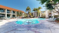 Located along Pacific Coast Highway in Torrance, California, this outdoor courtyard space offers a resort-inspired atmosphere defined by its freeform swimming pool and adjoining spa. The pool area is finished with brick-edged detailing and a spacious surrounding deck, providing ample room for lounge chairs and poolside relaxation under the Southern California sun. Towering palm trees and mature shade trees frame the space, creating a tropical ambiance that complements the warm coastal climate. Multiple seating areas with market umbrellas are arranged throughout the courtyard, offering shaded retreats for leisure. The complex's multi-story buildings, featuring warm stucco exteriors, green-accented balconies, and a distinctive red-tiled roof on one wing, encircle the courtyard, creating a sense of privacy and enclosure. A secondary, more intimate courtyard is also visible, featuring manicured garden beds with colorful plantings, decorative urns, and brick-accented pathways lined with iron railings and staircases. Lush greenery, including succulents, tropical foliage, and ornamental trees, is thoughtfully integrated throughout, enhancing the verdant character of the outdoor environment. Together, these spaces create a cohesive and inviting outdoor living experience ideal for the California lifestyle.