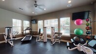 This single-level fitness center offers a comprehensive workout environment designed to accommodate a variety of exercise needs. The space is finished with durable dark rubber flooring throughout, providing a practical and slip-resistant surface suitable for both cardio and strength training activities. Walls feature a two-tone paint scheme with a warm brown lower section and neutral upper tone, complemented by white trim and baseboards.

Natural light enters generously through multiple white-slatted windows fitted with blinds, several of which overlook an outdoor pool area, adding a pleasant visual connection to the exterior. A ceiling fan with integrated lighting anchors the center of the room, supplemented by recessed lighting to ensure consistent illumination throughout.

The equipment selection includes treadmills, a stationary bike, and multiple selectorized strength training machines targeting various muscle groups, along with a medicine ball rack offering an assortment of weighted balls. Wall-mounted flat-screen televisions are positioned for visibility from cardio equipment. A glass-paned door provides access to an adjacent area. The overall layout allows for comfortable circulation between stations, supporting an efficient and functional fitness experience.