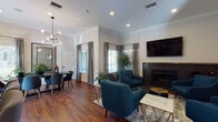 Located at Village Common in Fremont, CA, this single-floor community space combines contemporary design with inviting, functional interiors. Rich hardwood flooring flows throughout, anchoring the warm and cohesive aesthetic across each area.

The central lounge is a standout feature, arranged around a sleek dark-tiled fireplace topped with a wall-mounted flat-screen TV. A set of teal mid-century modern armchairs surrounds a marble-and-gold geometric coffee table, creating a comfortable and visually striking gathering area. Crown molding lines the ceilings, adding a refined architectural detail, while recessed lighting and multiple windows dressed in layered drapes and blinds allow for ample natural light.

Adjacent to the lounge, the dining area offers a large dark wood table surrounded by coordinating upholstered chairs beneath a sculptural globe chandelier. A decorative full-length mirror with a geometric lattice frame reflects the room and amplifies the sense of space, while abundant windows overlook the outdoor greenery.

The kitchen features warm wood cabinetry, light countertops, and a large black French door refrigerator, providing practical amenities for the space. French doors in the lounge area open to an outdoor area, further enhancing the connection between interior and exterior. Overall, the space presents a polished, welcoming environment well-suited for community gatherings.