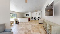 Located at 1250 S Euclid St in Anaheim, California, this single-floor space offers a well-organized and functional layout comprising a living room and an office area. The interior is defined by vaulted ceilings that lend an open, airy feel, complemented by multiple ceiling fans that promote airflow throughout. Large-format stone-look ceramic tile flooring extends seamlessly across the entire space, providing a clean, cohesive aesthetic.

The living room is generously proportioned, featuring wall-mounted flat-screen televisions, an ornate sideboard, and comfortable upholstered seating arranged to encourage relaxation and socializing. Natural light pours in through double glass entry doors at the front, offering views to the exterior and brightening the room. A decorative wall clock and framed artwork add character to the neutral white walls.

The office area is equipped with built-in desk workstations and ergonomic chairs, making it well-suited for computer use and administrative tasks. A wall-mounted air conditioning unit ensures climate control in this zone. An additional room serves as a flexible storage and multi-purpose space, currently housing folding chairs, tables, and miscellaneous items. A wet bar or kitchenette with cabinetry and a sink rounds out the utility of the space, supporting a variety of community or professional functions.