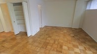 This single-floor space offers a clean, straightforward layout with a welcoming sense of openness. The most distinctive feature is the classic parquet wood flooring, laid in a traditional block pattern that adds warmth and character to the interior. The light-toned wood tiles create a visually rich surface underfoot, complementing the crisp, white-painted walls that keep the space feeling bright and airy.

The open layout allows for flexible use of the main area, which benefits from neutral wall tones that make it easy to personalize with furnishings and décor. A doorway leads to an adjacent space, supporting a functional flow between rooms. To one side, a built-in shelving unit provides practical storage, recessed neatly into the wall and framed by a white door, ideal for organization without sacrificing floor space.

The overall ambiance is understated and versatile — the combination of natural wood flooring and clean white surfaces creates a timeless, neutral aesthetic. The space is well-suited for a variety of uses and offers a solid foundation for customization. Its simple, uncluttered design ensures that the architectural details, particularly the parquet flooring, remain the standout element throughout.