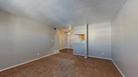 This single-floor unit presents a practical and well-organized layout across its living spaces. The living area is a generous, open room finished with wall-to-wall carpet in a warm tan tone, complemented by neutral-painted walls and a textured ceiling. A ceiling fan provides overhead comfort, while a pass-through opening connects the space to the kitchen, enhancing the sense of openness. Flooring transitions mark the boundary between the carpeted living space and the kitchen's durable hardwood-style flooring.

The galley-style kitchen is equipped with warm honey-toned wood cabinetry on both sides, light-colored countertops, and a tile backsplash with a subtle geometric pattern. Appliances include a white electric range and a stainless steel refrigerator, with a dishwasher also present. A flush-mount ceiling light illuminates the workspace efficiently.

The bedrooms are carpeted and finished with the same neutral palette, each featuring a window fitted with horizontal blinds that allow for natural light and privacy. The textured ceilings carry throughout these rooms, maintaining a consistent aesthetic.

The bathroom includes a full tub and shower combination with cream-toned tile surround, a toilet, and a vanity with a wood cabinet base. Hardwood-style flooring extends into this space, tying the wet areas together cohesively.