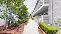 Located at 10740 Blix St in North Hollywood, Los Angeles, this single-level outdoor courtyard space serves as a welcoming common pathway connecting the residential buildings on the property. The corridor is defined by a wide, clean concrete walkway flanked on both sides by raised brick planter beds, which house a variety of well-maintained greenery including trimmed hedges, leafy trees, and ornamental shrubs. The plantings add a lush, verdant character to the passage, creating a pleasant garden-like atmosphere between the buildings.

The surrounding structures feature a consistent architectural palette of gray horizontal lap siding and textured stucco exteriors, giving the property a cohesive, modern-yet-classic aesthetic. Wall-mounted light fixtures are positioned along the building facades, ensuring the walkway is well-lit. At the far end of the courtyard, a set of white stairs with metal railings provides access to upper levels, while a glass-paneled entry door adds a refined touch to the building entrance. Additional seating and potted plants are visible toward the rear of the corridor, further enhancing the outdoor amenity space. The overall ambiance is clean, orderly, and thoughtfully landscaped, offering residents an attractive and functional shared outdoor environment.