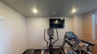 This single-floor fitness center offers a focused and functional workout environment designed to meet essential exercise needs. The space features a clean, minimalist aesthetic with light-toned walls and a smooth gray ceiling accented by recessed lighting, creating a well-lit and comfortable atmosphere for physical activity. The flooring is finished with dark rubber gym matting, providing both safety and durability underfoot. The room is equipped with key cardio machines, including an elliptical trainer and a treadmill, positioned to allow ease of use and adequate spacing between equipment. A wall-mounted flat-screen television is centrally positioned, providing entertainment during workouts. The recessed ceiling lights ensure even illumination throughout the space, while a smoke detector reflects attention to safety standards. The overall layout is efficient and straightforward, making the most of the available area without unnecessary clutter. The neutral color palette and practical design lend the space a professional yet approachable feel, suitable for a range of users. This fitness room provides a convenient, self-contained exercise facility with the essential amenities needed for a productive workout routine.