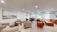 Located in Nashua, New Hampshire, this well-appointed single-floor space offers a thoughtfully designed combined dining and living area that balances comfort with functionality. The open layout is unified by light-toned hardwood-style flooring and crisp white walls, creating a clean, inviting atmosphere throughout.

The dining area features a rustic reclaimed-wood table paired with tufted dark leather chairs, offering a cozy setting for gatherings. Multiple windows dressed with horizontal blinds allow natural light to fill the space, with views of surrounding greenery. A decorative wall-mounted arrangement of sculptural metallic discs adds an artistic focal point, while a wall-mounted mini-split HVAC unit ensures year-round comfort.

The living area is furnished with a mix of tan upholstered club chairs, a classic leather sofa, and a solid wood coffee table, all anchored on a neutral area rug. Recessed ceiling lighting provides consistent illumination throughout. Framed abstract artwork adorns the walls, contributing to the space's refined yet approachable character. A wooden credenza along one wall adds practical storage and display space.

A secondary seating arrangement with upholstered armchairs centered around a natural wood stump-style side table offers a relaxed conversation nook. The space also includes access to a small kitchen area visible in the background, adding to its overall utility. Structural columns subtly define zones while maintaining the open feel of the floor plan.