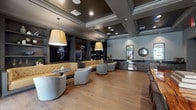 This upscale single-floor space blends a contemporary lounge aesthetic with functional meeting capabilities. The open layout is anchored by warm-toned hardwood flooring that flows throughout, complemented by a dark coffered ceiling with recessed lighting that lends an intimate, club-like ambiance. The lounge area serves as the centerpiece, featuring a tufted caramel leather banquette that wraps along the back wall, paired with round marble-top pedestal tables and curved gray upholstered barrel chairs. Oversized pendant lights in a muted yellow tone hang above the seating clusters, casting a warm glow. The back wall is finished in deep charcoal with built-in shelving and dramatic large-format black-and-white art prints, adding a bold artistic statement. Adjacent to the lounge is a wet bar area equipped with dark veined stone countertops, gray cabinetry, a stainless steel under-counter refrigerator, a microwave, and a black matte sink — ideal for entertaining. A sunburst decorative mirror adds a sculptural accent above the bar. The meeting area features a striking live-edge wood conference table surrounded by upholstered side chairs, with a wall-mounted display screen positioned for presentations. Large windows allow natural light to filter into the space, balancing the richly layered interior palette.