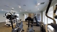 This fitness center is thoughtfully designed to support a well-rounded workout experience. The space is outfitted with a comprehensive selection of cardio equipment, including stationary bikes, elliptical machines, treadmills, and stair climbers, providing members with a wide range of aerobic training options. Along the mirrored wall, strength training equipment and free weights offer additional versatility for resistance-based workouts. The layout is organized to allow comfortable movement between machines, making efficient use of the available floor space. Underfoot, durable carpet provides cushioning throughout the workout area. The room is illuminated by recessed ceiling lights that offer consistent, even brightness at all hours. Large windows fitted with horizontal blinds line the walls, inviting ample natural light and allowing for ventilation while maintaining privacy. The textured ceiling and sage-green walls contribute to a calm, focused atmosphere conducive to exercise. A wall-mounted light fixture and in-ceiling speakers add functional and ambient touches to the overall environment. The single-floor layout ensures easy accessibility, making this fitness center a practical and well-appointed space for daily exercise routines.