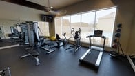 This single-level fitness center is designed to provide a functional and well-equipped workout environment. Large floor-to-ceiling windows span one wall, flooding the space with natural light and offering views of an outdoor pool area, creating an open and motivating atmosphere. The rubber flooring throughout the room provides durability and safety for a variety of exercises.

The cardio section is equipped with a treadmill, elliptical trainer, and a recumbent bike, offering multiple options for aerobic workouts. A cable weight machine and a weight bench with a stability ball round out the strength training area, providing a versatile range of resistance exercises. A wall-mounted mirror expands the sense of space and allows users to monitor their form during workouts. A flat-screen television mounted on the wall adds entertainment value during exercise sessions.

A ceiling fan ensures comfortable air circulation throughout the room. The neutral-toned walls and dark flooring create a clean, focused aesthetic. Overall, the space is efficiently arranged to maximize usability while maintaining an uncluttered and welcoming workout environment.