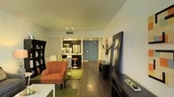 This single-floor space is designed with an open-concept layout that connects the living area and kitchen, creating an efficient and cohesive flow. The living area features rich dark hardwood flooring that extends throughout, grounding the space with warmth and contrast against the light-toned walls and ceiling. Recessed and track lighting provides well-distributed illumination across the main areas.

The living room is anchored by a bold rust-orange armchair, complemented by a green area rug that adds a pop of color and texture underfoot. A large decorative mirror leans against one wall, enhancing the sense of depth and light, while a tall bookshelf along the adjacent wall offers storage and display space. A low media console anchors the entertainment area, keeping the layout practical and organized.

The kitchen is tucked neatly toward the rear, featuring dark upper cabinetry, stainless steel appliances including a refrigerator and microwave, and a raised breakfast bar with seating. A barstool at the counter reinforces the casual dining option. The entry door, finished in a slate-blue tone, adds a subtle design accent. Overall, the space maintains a modern, urban aesthetic with thoughtful use of color and furnishings.