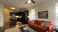 This single-floor residence offers an open-concept layout that seamlessly blends the living and kitchen areas into one cohesive, functional space. The living area is warm and inviting, featuring rich hardwood flooring that extends throughout, a ceiling fan with integrated lighting, and a bright window dressed with sheer white curtains and blinds that allow natural light to fill the room. The kitchen is a standout feature, showcasing bold dark cabinetry that reaches toward the ceiling, black appliances including a full-size refrigerator and microwave, a kitchen island that provides additional prep and gathering space, and under-cabinet lighting that adds both practicality and ambiance. The contrast between the dark cabinetry and the light-toned walls creates a striking visual balance throughout the open space. The living area comfortably accommodates a full sofa and coffee table arrangement, making it well-suited for both relaxing and entertaining. The overall design aesthetic is modern and cohesive, with a consistent use of dark tones and warm wood accents that lend the space a grounded, stylish character.