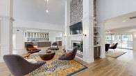 Located at 450 Technology Pkwy NW in Norcross, Georgia, this beautifully appointed single-floor clubhouse offers a refined and welcoming atmosphere designed for community gathering and everyday amenity use. The open-concept layout is anchored by a dramatic floor-to-ceiling stone fireplace flanked by white-paneled columns and built-in shelving, creating a striking focal point throughout the main lounge area. Soaring ceilings with clerestory windows flood the interiors with abundant natural light, complementing the warm-toned hardwood flooring that flows seamlessly throughout the space. The lounge is furnished with contemporary seating arrangements atop vibrant area rugs, fostering an inviting environment for relaxation or socializing. A dedicated workspace area with desks and glass-partitioned offices provides a functional setting for remote work or meetings. The expansive kitchen features rich dark wood cabinetry, white quartz countertops, a large central island, glass-front upper cabinets, a herringbone tile backsplash, and statement pendant lighting. A bright, open event room with direct access to the outdoor pool area through French doors offers versatile space for gatherings. A fitness center with mirrored walls and modern equipment is also visible, rounding out this well-equipped community facility.