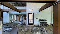 This fitness center offers a comprehensive workout environment designed to accommodate a wide range of exercise routines. The space features soaring ceilings with exposed ductwork and bold dark ceiling beams that lend an industrial-modern character to the interior. Large windows along one wall allow natural light to fill the space, creating an open and energizing atmosphere.

The workout floor is equipped with a full dumbbell rack offering a complete range of free weights, along with a dedicated adjustable weight bench for strength training. A multi-use cable and strength machine is positioned toward the center of the space, providing options for a variety of resistance exercises. Rubber floor mats are laid throughout key workout zones to provide stability and protect the flooring during high-impact use.

The flooring consists of a patterned carpet in the surrounding areas, transitioning to darker rubber matting in active training zones. A glass-paneled exterior door provides direct outdoor access, adding ventilation options and a connection to the outside. The neutral, light-toned walls keep the environment feeling clean and focused. Overall, the fitness center is thoughtfully laid out to support both strength and functional training within a bright, well-maintained facility.