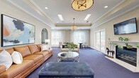 Located at 4835 Cameron Valley Parkway in Charlotte, NC, this well-appointed community gathering space offers a refined and welcoming atmosphere designed for both relaxation and casual meetings. The main lounge area makes an immediate impression with its tray ceiling adorned with stylish pendant lighting and recessed fixtures, alongside warm beige walls and rich blue carpeting that flows throughout the space. A leather sofa, upholstered seating, and a round glass coffee table form an inviting conversation area, while a wall-mounted fireplace with a classic white mantel and flat-screen TV anchors one end of the room. Multiple sets of French doors open to an outdoor patio, flooding the interior with natural light and providing seamless indoor-outdoor connectivity. A separate dining or meeting area features round pedestal tables paired with neutral upholstered chairs, positioned beside large windows dressed with sheer curtains and wood blinds that frame views of the surrounding greenery. A compact kitchenette with white cabinetry, a tile backsplash, and a sink offers convenient amenities for gatherings. Crown molding, arched mirrors, and colorful artwork contribute to the space's polished, community-oriented aesthetic throughout.