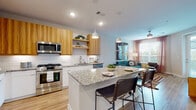 This single-floor residence offers a thoughtfully designed open-concept layout where the kitchen and living area flow seamlessly together. The kitchen is a standout feature, combining white lower cabinetry with warm wood-toned upper cabinets and open shelving. Granite countertops extend across the perimeter and onto a central island, which provides additional seating and prep space. Stainless steel appliances, including a gas range, microwave, dishwasher, and French door refrigerator, complete the modern setup. White subway tile backsplash adds a clean, classic contrast to the wood accents, while pendant lighting above the island enhances the functional aesthetic.

The living area benefits from multiple windows that allow generous natural light to fill the space. A ceiling fan overhead and a direct exterior door contribute to the room's practical appeal. Light wood-style flooring runs consistently throughout the entire unit, unifying each space visually.

The bathroom features a full tub-shower combination with large-format tile surround, a white vanity with granite countertop, and built-in open shelving for additional storage. The walk-in closet provides organized wire shelving systems on multiple walls, offering ample hanging and storage capacity throughout.