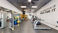 This fitness studio is housed within a single-floor space that makes efficient use of its open layout to accommodate a comprehensive range of workout equipment. The interior is designed with a clean, high-energy aesthetic, featuring white walls adorned with bold motivational typography — including phrases such as "Train Hard," "Strength," and "Don't Wish for It, Work for It" — that reinforce the studio's active atmosphere.

The ceiling is finished in dark paneling with recessed fluorescent lighting and ceiling fans, contributing to a focused, gym-style ambiance. The flooring throughout consists of durable rubber matting, practical for both heavy equipment and high-impact exercise.

One full wall features a distinctive horizontal wood slat installation, which serves as an organizational and display surface for accessories such as medicine balls, resistance bands, and other functional training tools. A full dumbbell rack is positioned nearby, offering a range of free weights. On the opposite side of the studio, a row of cardio machines — including treadmills and ellipticals — lines the space, facing a wall-mounted television.

Additional equipment includes weight benches, a leg extension machine, a cable/pulley system, stability balls, and a mirror wall that enhances the sense of space. A glass-panel entry door at one end allows natural light into the studio.
