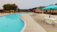This outdoor pool area serves as a central recreational amenity within the community. The freeform pool is notably large, featuring gently curved edges and vivid turquoise water with clearly marked depth indicators along the pool's edge. Entry is provided via built-in steps with decorative tiled detailing, as well as metal handrail access points along the pool's perimeter.

The surrounding concrete deck is spacious, offering ample room for movement and relaxation. A generous row of white lounge chairs lines one side of the pool, providing dedicated sunbathing space. Multiple seating areas are positioned throughout the deck, each featuring a round table accompanied by chairs and shaded by brightly colored market umbrellas in yellow, red, and teal tones, creating a lively, resort-like atmosphere.

The pool area is enclosed by a black wrought iron fence, providing security while maintaining open sightlines to the surrounding landscaping. Mature deciduous trees border the perimeter, offering a natural backdrop. A brick and siding accessory building is situated adjacent to the deck, likely serving as a pool house or utility structure. The overall setting is open, functional, and well-equipped for outdoor leisure.