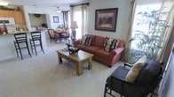 This single-story space showcases an open-concept layout that seamlessly connects the living area to the dining space, creating a cohesive and functional floor plan. The living room is anchored by a plush, rust-colored sofa accented with decorative floral and patterned throw pillows, complemented by a rustic wooden coffee table with ornamental detailing that adds a touch of character to the room. A coordinating seating arrangement is partially visible, contributing to a comfortable, conversational setup. Framed wall art adds a polished finishing touch above the sofa. Soft, neutral carpeting runs throughout the space, enhancing the warm and inviting ambiance. Natural light filters in generously through multiple windows dressed with layered curtains, brightening the interior throughout the day. A floor lamp provides additional ambient lighting. The adjacent dining area accommodates a round table surrounded by cushioned chairs, ideal for everyday meals or entertaining. A glimpse into what appears to be a kitchen is visible beyond the dining space, suggesting a practical and connected layout. Overall, the space presents a traditional aesthetic with warm tones, comfortable furnishings, and a well-considered flow between living areas.
