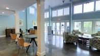 This single-story space makes a strong architectural impression through its soaring double-height ceilings, which are framed by rows of clerestory windows that flood the interior with natural light. The walls are painted in a calming light blue-gray tone, complementing the highly polished concrete floors that reflect light throughout the open layout.

Structural white columns are positioned throughout the space, adding visual interest while defining distinct zones within the open floor plan. A seating area near the entrance features patterned upholstered armchairs, providing a welcoming lounge setting. A small café-style table with bentwood chairs occupies a separate area to one side, suggesting an informal gathering or work spot.

A reception or administrative desk area is visible toward the rear left, indicating this space likely functions as a communal lobby or clubhouse. Large glass French doors at the entry allow views to an outdoor area beyond, enhancing the connection between interior and exterior. Pendant lights and ceiling fans hang from above, contributing both functionality and a clean, contemporary aesthetic. Overall, the space conveys an airy, well-lit, and inviting atmosphere.