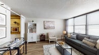 This single-floor residence offers a thoughtfully designed layout that balances comfort and functionality across its key living spaces. The living room serves as the heart of the unit, featuring warm hardwood-style flooring, neutral walls, and a textured ceiling throughout. A bank of windows dressed with sheer curtains and horizontal blinds floods the space with natural light while framing open exterior views. The room accommodates both a seating area anchored by a gray sofa and a dedicated reading nook with a classic upholstered accent chair beneath a large abstract canvas.

The galley-style kitchen is efficiently arranged with white cabinetry, granite-look countertops on both sides, a white gas range, and hardwood-style flooring that continues throughout. A louvered closet door provides additional storage along the corridor.

The bedroom is a tranquil retreat featuring corner windows that offer sweeping panoramic views of the surrounding cityscape. The hardwood-style floors continue here, complemented by neutral tones and soft, layered bedding.

The bathroom is finished with mosaic tile flooring, cream wall tiles, a white vanity with granite countertops, and a full tub-shower combination, providing a clean and functional space.