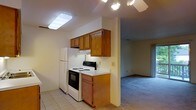 This single-floor space offers a practical and straightforward layout, with a kitchen that flows openly into an adjacent living area. The kitchen is equipped with essential appliances, including a white refrigerator and an electric range with coil burners, complemented by a built-in range hood above. Warm-toned medium wood cabinetry lines the upper walls, providing ample storage space, while light-colored countertops offer a functional work surface. The kitchen floor is finished in neutral light tile, providing a clean and easy-to-maintain surface. Overhead, a flush-mount light fixture ensures adequate task lighting throughout the cooking area. The adjacent living space transitions seamlessly from the kitchen, featuring wall-to-wall carpeting in a neutral tone that adds warmth underfoot. A ceiling fan with an integrated light kit serves this area, offering both airflow and ambient illumination. The overall color palette throughout the space is light and neutral, with cream-toned walls contributing to a bright, open feel. The layout is efficient and functional, making practical use of the available space across the single floor.