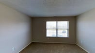 This single-story space presents a clean, functional layout designed for comfortable everyday living. The room pictured features wall-to-wall carpeting in a warm neutral tone, providing a soft and practical flooring surface throughout. The walls are finished in a understated beige, creating a versatile backdrop that pairs well with a variety of furnishing styles. A prominent double-pane window fitted with horizontal blinds serves as a key architectural feature, allowing natural light to fill the room while offering a view of the outdoor surroundings, including greenery and a paved area beyond. The textured popcorn ceiling adds a classic touch consistent with traditional residential construction. A visible HVAC vent integrated into the ceiling indicates a central climate control system. The baseboard trim runs cleanly along the perimeter of the room, adding a finished, polished look to the overall interior. The space is presented in vacant condition, making it easy to envision various furniture arrangements and uses. Overall, the room conveys a straightforward, move-in-ready aesthetic suited to a range of occupants.