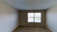 This single-story space presents a clean, functional layout designed for comfortable everyday living. The room pictured features wall-to-wall carpeting in a warm neutral tone, providing a soft and practical flooring surface throughout. The walls are finished in a understated beige, creating a versatile backdrop that pairs well with a variety of furnishing styles. A prominent double-pane window fitted with horizontal blinds serves as a key architectural feature, allowing natural light to fill the room while offering a view of the outdoor surroundings, including greenery and a paved area beyond. The textured popcorn ceiling adds a classic touch consistent with traditional residential construction. A visible HVAC vent integrated into the ceiling indicates a central climate control system. The baseboard trim runs cleanly along the perimeter of the room, adding a finished, polished look to the overall interior. The space is presented in vacant condition, making it easy to envision various furniture arrangements and uses. Overall, the room conveys a straightforward, move-in-ready aesthetic suited to a range of occupants.