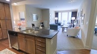 This single-floor residence is thoughtfully designed around an open-concept layout that connects the kitchen, dining, and living areas into one cohesive, functional space. The kitchen anchors the layout with a prominent island topped with dark granite countertops, a double stainless steel sink, and warm wood cabinetry featuring brushed nickel hardware — combining both style and practicality.

The kitchen opens directly into a casual dining area, which features a glass-top table and dark chairs beneath a wall-mounted decorative mirror, creating an intimate yet elegant setting. Beyond, the living room enjoys an abundance of natural light streaming through large windows dressed with dramatic dark curtains, lending contrast and sophistication to the neutral-toned walls.

Hardwood flooring runs throughout, grounding the space with warmth, while a soft area rug in the living room adds texture and definition. The living area comfortably accommodates a full seating arrangement, reinforcing the spacious feel of the open floor plan. Overall, the space strikes a balance between modern design and inviting comfort.