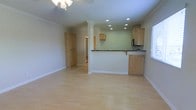 This single-floor space offers an open, airy layout that combines the living and kitchen areas into one cohesive environment. Light hardwood flooring runs throughout, contributing to a warm and welcoming ambiance that reflects natural light from a window along the side wall. The living area is generously sized, with crown molding along the ceiling and a ceiling fan with light fixture providing both comfort and style. Recessed lighting is thoughtfully installed throughout, offering bright, even illumination across the space. The kitchen is defined by a raised breakfast bar with a dark countertop that provides a natural visual boundary between the cooking and living zones. Light maple cabinetry lines the kitchen walls, offering ample storage, while a built-in microwave and sink add practical functionality. A column detail near the kitchen entry adds an architectural touch that subtly frames the transition between spaces. Adjacent to the main living area, a hallway leads to additional rooms, with a full-height storage closet visible nearby. The overall palette — soft green walls, white trim, and natural wood tones — creates a calm, neutral aesthetic that is both functional and inviting.