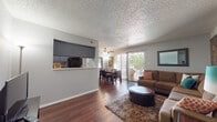 This single-floor residence offers a thoughtfully designed open-concept layout that seamlessly connects the living, dining, and kitchen areas. Rich hardwood flooring extends throughout the main living spaces, complemented by a textured ceiling and neutral gray walls that create a clean, cohesive aesthetic.

The spacious living room is anchored by a large sectional sofa and features a sliding glass door that opens to a private balcony overlooking greenery, allowing ample natural light to fill the space. A ceiling fan adds both function and style to the room.

Adjacent to the living area, the dining space accommodates a full dining table and chairs, with vertical blinds on the sliding door providing flexible light control. The pass-through window connects the dining area to the galley-style kitchen, which features dark cabinetry, white countertops, a black electric range, and an overhead exhaust hood — offering an efficient and practical cooking space.

The bedroom is carpeted for comfort and includes a built-in closet with shelving, providing generous storage. It connects directly to a private bathroom, which features a wide vanity with light countertops, dark cabinetry, hardwood flooring, and a large mirror. The overall design balances functionality with a welcoming residential atmosphere throughout.