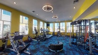 This single-floor fitness center is designed to offer a comprehensive workout experience in a bright, energetic environment. The space is immediately distinguished by its bold yellow walls and vivid blue plaid carpeting, creating a lively and motivating atmosphere throughout. Large windows line multiple walls, flooding the interior with natural light and providing open views of the surrounding outdoor environment, enhancing the sense of space and airiness.

The fitness center is generously outfitted with a diverse selection of equipment to support a full range of training disciplines. A dedicated cardio zone features multiple treadmills, elliptical machines, and stationary bikes, all positioned near the windows to allow users to exercise with an outdoor view. Overhead, mounted flat-screen televisions provide entertainment during workouts.

The strength training area is equally well-appointed, including a functional training rig with resistance attachments, a tiered dumbbell rack, a weight bench, and a variety of cable and plate-loaded machines. A large tractor tire and sled-style equipment signal dedicated space for functional and high-intensity training.

Distinctive circular pendant light fixtures with integrated fans hang from the white ceiling, complementing the recessed lighting throughout. The overall design balances energetic aesthetics with practical, professional-grade fitness amenities.