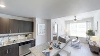 This single-floor residence showcases a thoughtfully designed open-concept layout that seamlessly connects the kitchen, dining, and living areas. The kitchen is equipped with dark cabinetry contrasted by light granite countertops, a mosaic tile backsplash, and stainless steel appliances, including a dishwasher. A kitchen peninsula doubles as a dining surface, offering casual seating and additional workspace.

The dining and living areas flow naturally from the kitchen, creating a cohesive and functional shared space. The living room benefits from abundant natural light streaming through large windows dressed with patterned curtains, and is equipped with a ceiling fan for added comfort. Light-toned flooring runs throughout, enhancing the airy, open feel of the space.

A glimpse into the adjacent bedroom reveals a bright, neatly appointed room with its own framed wall art and access to additional areas of the unit. Neutral walls throughout provide a clean, versatile backdrop. The overall ambiance is modern and inviting, with a consistent color palette of whites, grays, and dark accents lending the space a polished, contemporary character.