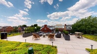 This rooftop terrace is a thoughtfully designed outdoor space that combines sustainability with comfort and style. A defining feature of the terrace is its extensive green roof, which is blanketed in lush, low-growing sedum and other ground cover plants that provide both visual appeal and environmental benefit. The living greenery stretches across a significant portion of the roof surface, contrasting beautifully with the sleek, dark-paneled architectural facade of the building's rooftop structure.

The terrace includes multiple distinct lounge areas furnished with woven rattan and upholstered seating, coffee tables, and accent chairs, creating inviting spaces suited for both relaxed gatherings and quiet outdoor enjoyment. Large planters filled with manicured shrubs serve as natural dividers throughout the space, adding greenery and structure to the layout.

Underfoot, clean large-format stone pavers provide a refined, durable surface that ties the seating areas together. The perimeter is enclosed by a black metal railing, offering safety while preserving unobstructed sightlines to the surrounding neighborhood rooftops and mature tree canopy. Floor-level path lighting adds functionality for evening use.

Access to the terrace is provided through full-height glass and dark metal-framed doors, reinforcing the modern architectural language of the building. The open sky above and panoramic urban views make this rooftop an exceptional outdoor amenity.