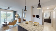 This single-floor residence showcases a thoughtfully designed open-concept layout that seamlessly connects the kitchen, dining, and living areas. The kitchen is a standout feature, equipped with stainless steel appliances including a French door refrigerator, electric range, built-in microwave, and dishwasher. Two-toned cabinetry pairs dark espresso lower cabinets with crisp white upper cabinets, accented by a classic subway tile backsplash. A granite-topped island with bar seating serves as a central gathering point, illuminated by striking black conical pendant lights.

The living area maintains a calm, neutral palette with soft sage-toned walls and teal drapery that frame a window offering views of lush greenery. Light tile flooring runs throughout the space, enhancing the bright and airy atmosphere.

Two bedrooms provide comfortable retreats, each featuring ceiling fans, natural light from windows with tropical foliage views, and botanical-patterned curtains that reinforce the space's relaxed aesthetic. The primary bedroom includes floating nightstands and mirror accents for a polished look.

The bathroom features full-height linear tile walls, a walk-in glass shower with a built-in bench, a granite vanity countertop, dark cabinetry, and a backlit LED mirror, delivering a clean, spa-inspired finish.