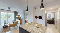 This single-floor residence showcases a thoughtfully designed open-concept layout that seamlessly connects the kitchen, dining, and living areas. The kitchen is a standout feature, equipped with stainless steel appliances including a French door refrigerator, electric range, built-in microwave, and dishwasher. Two-toned cabinetry pairs dark espresso lower cabinets with crisp white upper cabinets, accented by a classic subway tile backsplash. A granite-topped island with bar seating serves as a central gathering point, illuminated by striking black conical pendant lights.

The living area maintains a calm, neutral palette with soft sage-toned walls and teal drapery that frame a window offering views of lush greenery. Light tile flooring runs throughout the space, enhancing the bright and airy atmosphere.

Two bedrooms provide comfortable retreats, each featuring ceiling fans, natural light from windows with tropical foliage views, and botanical-patterned curtains that reinforce the space's relaxed aesthetic. The primary bedroom includes floating nightstands and mirror accents for a polished look.

The bathroom features full-height linear tile walls, a walk-in glass shower with a built-in bench, a granite vanity countertop, dark cabinetry, and a backlit LED mirror, delivering a clean, spa-inspired finish.