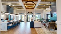 Located at 1020 Port Royal Trail in Myrtle Beach, SC, this single-story community clubhouse delivers an impressive blend of modern farmhouse aesthetics and resort-style amenities across a generously sized floor plan. The centerpiece is a dramatic open-concept great room showcasing soaring ceilings adorned with exposed rustic wood beams, a statement ring chandelier in brushed gold, and large black-framed windows that flood the space with natural light and offer views of the surrounding landscape. A sleek, full-length kitchen anchors one side of the room, featuring navy cabinetry, white quartz countertops with a bold marble-look backsplash, floating wood shelves, and globe pendant lighting over a spacious island with bar seating. A dedicated lounge area includes a shiplap accent wall with an integrated linear fireplace and mounted television, creating an inviting gathering spot. The fitness wing is thoroughly equipped with cardio machines, a multi-function cable system, free weights, medicine balls, and a separate strength and flexibility room with herringbone-style flooring. A private indoor golf simulator room rounds out the amenity offerings, complete with a putting green surface, spectator seating, and large windows overlooking the outdoors. Three connecting hallways link the functional zones seamlessly throughout the building.