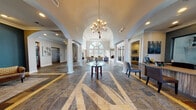 This single-floor clubhouse is defined by its impressive architectural design and refined interior finishes. The grand entrance lobby sets a distinguished tone with soaring vaulted ceilings, a stately arched window that floods the space with natural light, and pairs of fluted white columns framing elegant arched passageways. A decorative glass-top table anchors the center of the lobby beneath a brass chandelier, while slate-tile flooring with inlaid geometric border detailing extends throughout much of the space. A staffed reception desk is conveniently positioned near the entry, and an elevator provides additional accessibility.

Beyond the lobby, a spacious lounge area features double-height ceilings, floor-to-ceiling windows with outdoor views, a built-in media center, and comfortable seating arrangements bathed in abundant natural light. A casual dining and lounge area with high-top tables, leather seating, a wall-mounted television, and a ceiling fan offers a relaxed gathering environment. A well-appointed conference room includes wainscoted walls in a rich blue tone, a large wooden conference table, and refined crown molding. The fully equipped kitchen features white cabinetry, slate-tile backsplash, granite countertops, and full appliances, supporting events and everyday use.