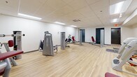 Located at 319 N Pottstown Pike in Exton, PA, this single-floor fitness facility offers a focused and functional workout environment across a compact yet efficiently organized footprint. The space is divided into two interconnected areas, both finished with light hardwood-style flooring that creates a clean, energetic atmosphere throughout.

The facility is outfitted with a comprehensive selection of professional-grade strength training equipment from well-known brands including Freemotion, Precor, and Matrix, featuring machines with distinctive silver frames and red upholstered seating. Equipment is thoughtfully arranged to allow comfortable movement between stations, targeting a wide range of muscle groups for a full-body workout experience.

Overhead, a drop-panel ceiling with recessed and flush-mounted fluorescent lighting provides consistent, even illumination across the entire space. Climate control is handled by ceiling-mounted HVAC units, ensuring a comfortable environment year-round. The walls are finished in a crisp white, contributing to a bright and open feel. Gray accent area rugs near the entryway add a subtle design touch and practical function.

Access is provided through two teal-painted doors, with clearly marked exit signage ensuring safety compliance. The overall design is clean, modern, and purposeful, making this facility a practical and well-appointed space for strength and fitness training.
