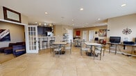 Located at 4175 Harding Hwy in Lima, Ohio, this single-floor community clubhouse offers a well-appointed gathering space designed to serve residents across a range of daily needs and social activities. The heart of the facility is a generously sized common dining and gathering area, finished with large-format neutral tile flooring and recessed lighting throughout. Multiple dining tables with metal-framed chairs are arranged throughout the open layout, providing ample seating for residents to gather, dine, or socialize. A full kitchen anchors one end of the main space, featuring dark cabinetry, a breakfast bar with bar-height seating, stainless steel appliances, and a white refrigerator. An adjacent prep kitchen or kitchenette offers additional counter space with dark cabinetry, a stainless steel sink, a dishwasher, and a coffee station. Natural light filters into the dining area through several windows with blinds, enhancing the bright and welcoming atmosphere. A dedicated lounge area provides a comfortable sectional sofa and wall-mounted television for relaxed viewing. Computer workstations are available along one wall for resident use. The facility also includes a well-equipped fitness center with a bold red and blue color scheme, housing a variety of cardio and strength training machines. A small seating nook near the entrance, complete with framed artwork and a patterned area rug, offers a welcoming reception area.