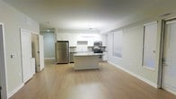 This single-floor space showcases a clean, modern design centered around an open-concept layout that seamlessly connects the kitchen and living areas. The kitchen is the focal point of the space, outfitted with stainless steel appliances including a full-size refrigerator, microwave, and range. Crisp white cabinetry lines the walls, complemented by a subway tile backsplash that adds a classic yet contemporary touch. A central island provides additional counter space and serves as a natural gathering point within the open layout. The countertops offer ample workspace, and under-cabinet areas are thoughtfully designed for both function and aesthetics. Track lighting overhead ensures bright, even illumination throughout the kitchen, while recessed lighting extends into the adjacent living area. Light hardwood flooring runs continuously across the space, enhancing the sense of openness and contributing to the unit's airy, welcoming ambiance. Neutral wall tones in soft gray and white create a versatile backdrop that complements a variety of décor styles. A window with blinds allows for natural light and privacy control. The overall design is cohesive, practical, and well-suited for modern living.